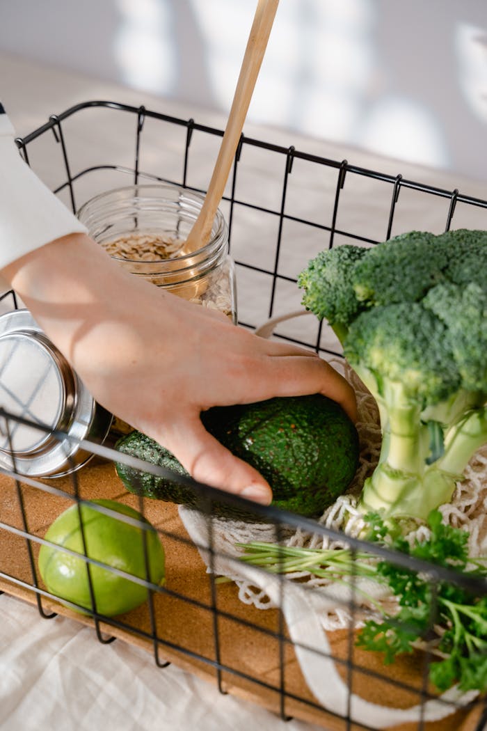 Hand reaches for avocado amidst broccoli and grains in wire basket for healthy cooking.